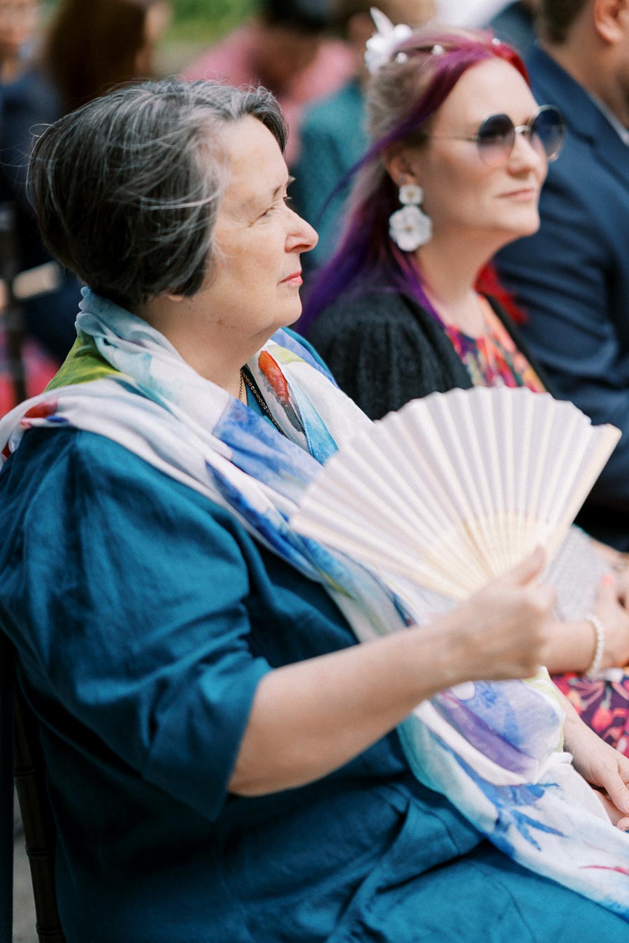 Elegant woman in a blue dress sitting at an outdoor event, holding a fan, with another person beside her wearing sunglasses and a floral outfit.