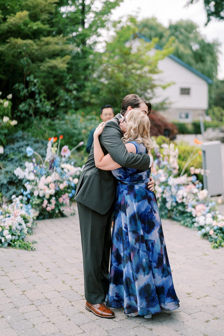Two people embracing at an outdoor event, surrounded by lush greenery and colorful flowers, with a focus on a heartfelt moment.