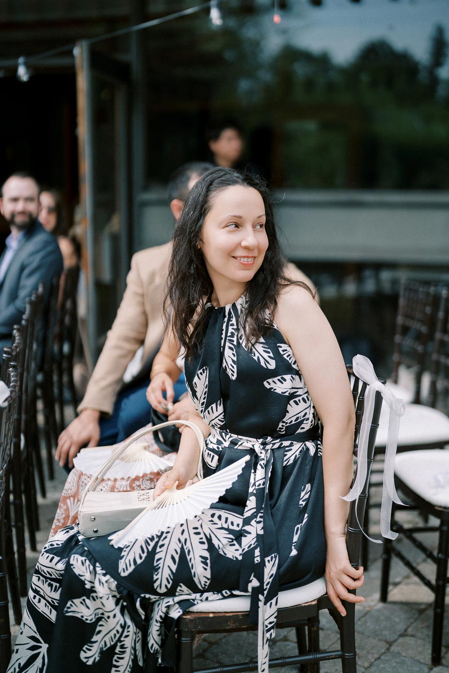 A woman in a patterned dress smiles while sitting outdoors at an event.