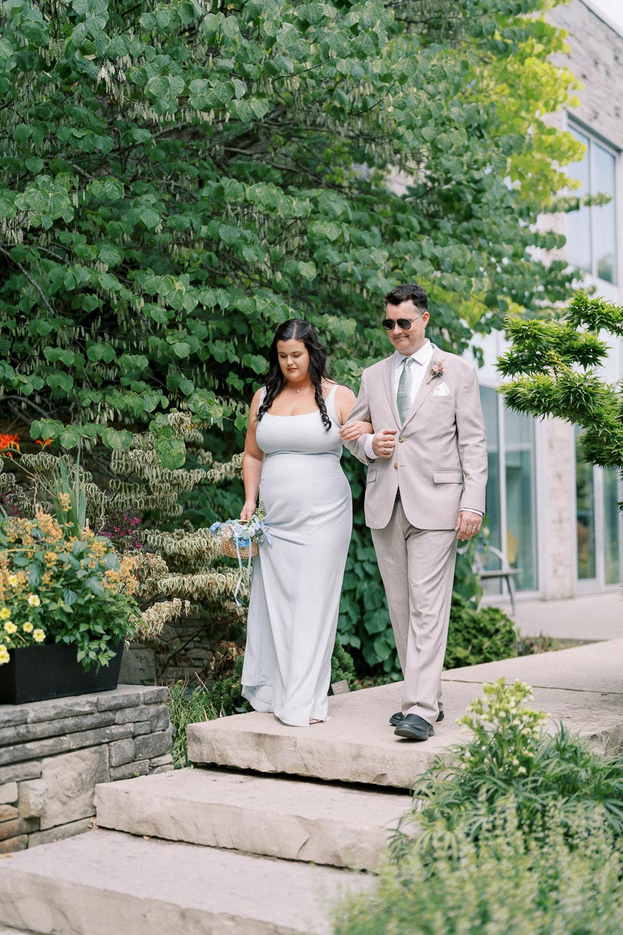 A bridesmaid in a light blue gown walks with a groomsman in a light gray suit down an outdoor stone pathway, surrounded by lush greenery and vibrant flowers.
