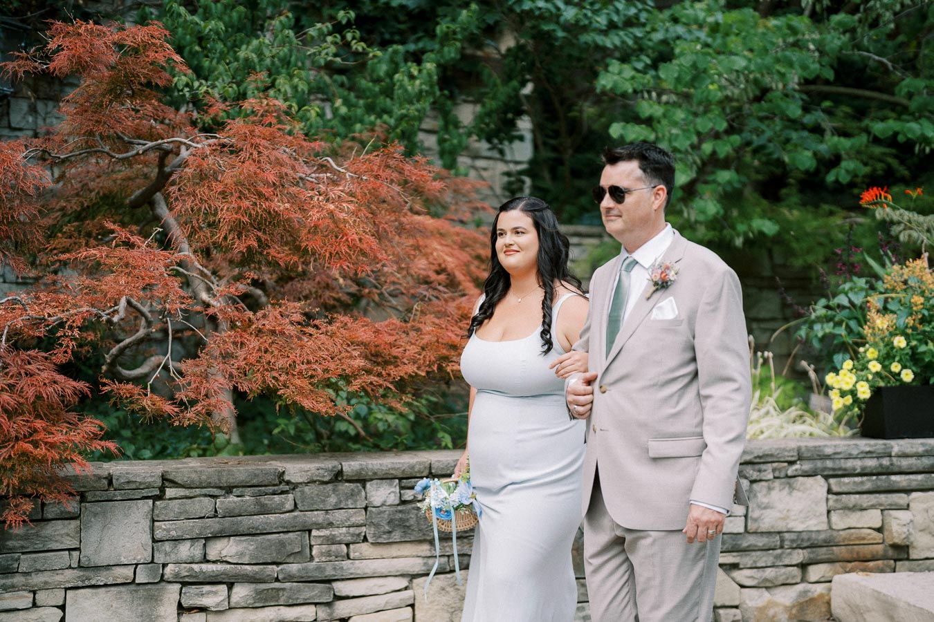 A woman in a light gray dress and a man in a beige suit walking arm in arm down a stone path, surrounded by lush greenery and red foliage, with decorative flowers nearby.