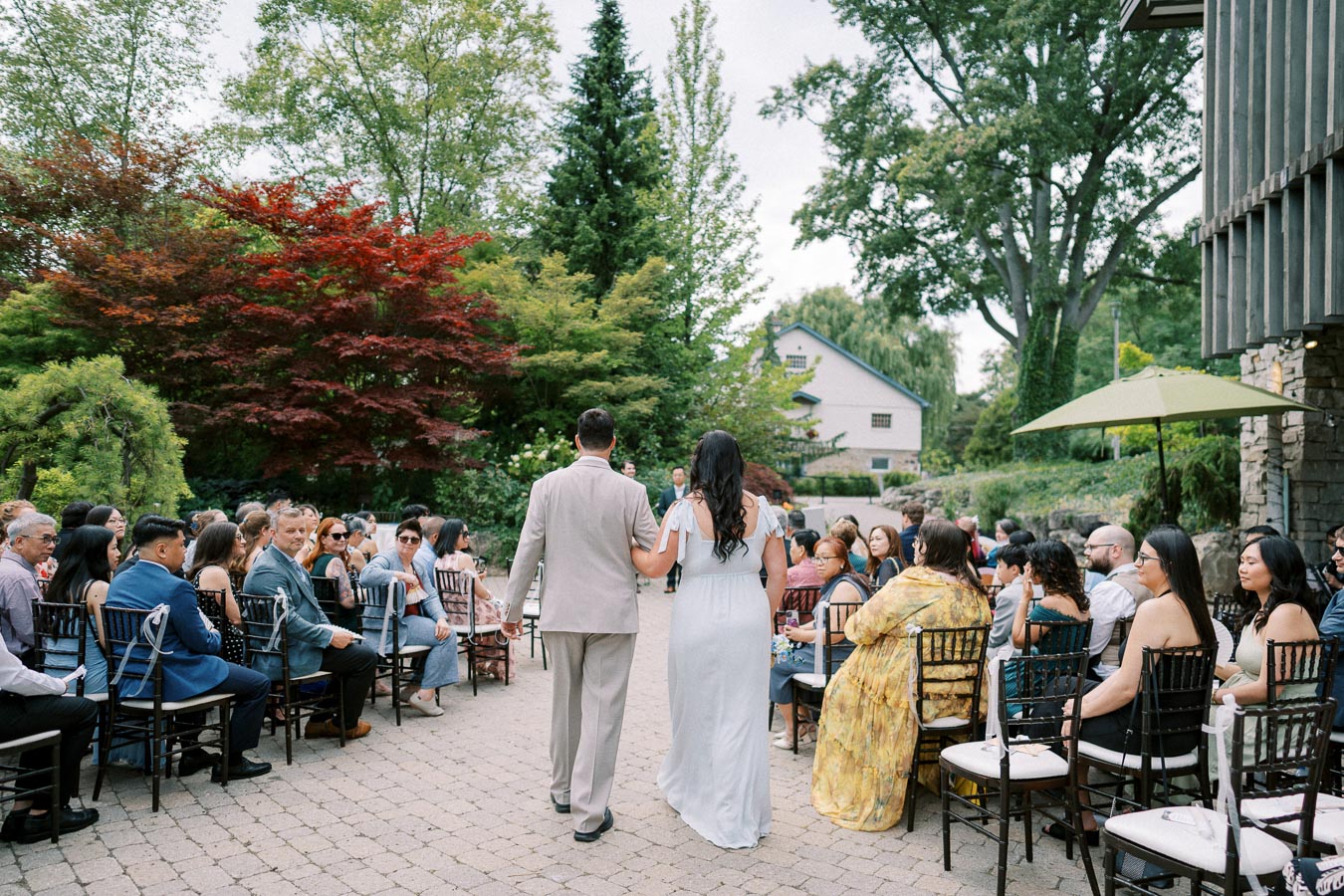 Outdoor wedding ceremony with couple walking down the aisle, surrounded by guests seated on either side, lush greenery and trees in the background.
