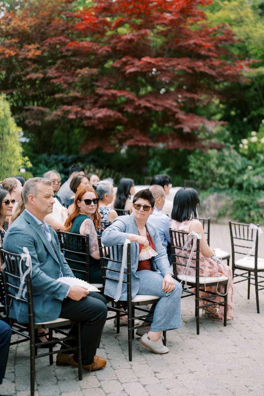 A stylish group of wedding guests seated outdoors with lush greenery and red-leafed trees in the background, creating a serene atmosphere.