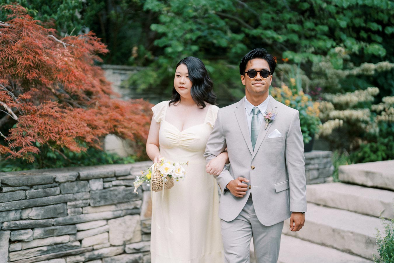 A couple walking arm in arm at an outdoor wedding ceremony, with the woman in a pale yellow dress holding a flower bouquet, and the man in a light gray suit with a teal tie, sunglasses, and a boutonniere. They are surrounded by lush greenery and a stone wall backdrop.