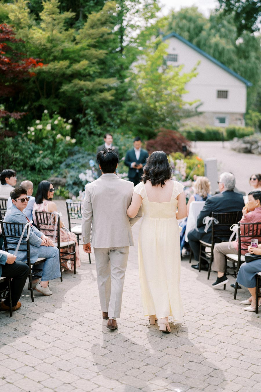 Outdoor wedding ceremony with a couple walking down the aisle, surrounded by seated guests in elegant attire, lush greenery in the background creating a serene and picturesque setting.