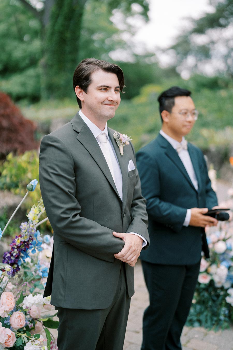 Man in a green suit smiling at an outdoor wedding ceremony, surrounded by lush greenery and colorful flowers.