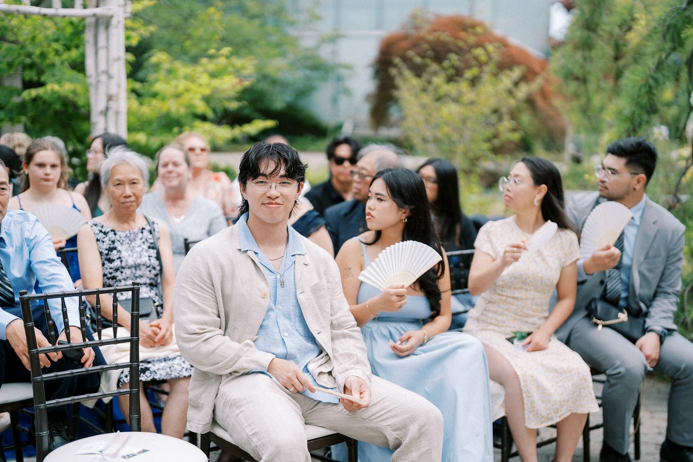 A diverse group of well-dressed people sitting outdoors at a formal event. The individuals appear engaged and are fanning themselves with hand fans. The setting is lush and green, indicating a possible summer gathering or celebration.