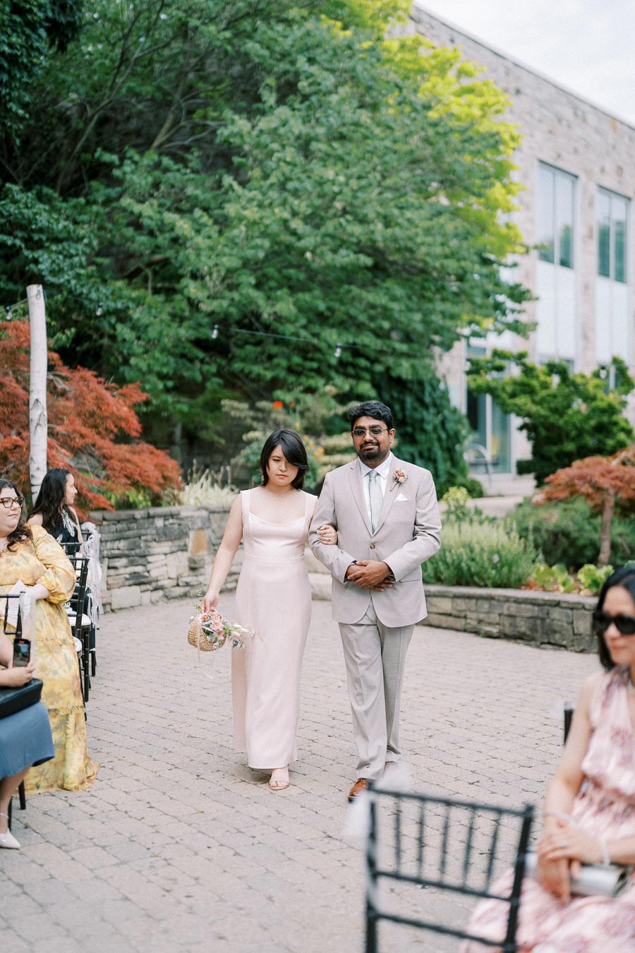 Elegant outdoor wedding procession with a woman in a flowing pink dress holding a basket of flowers, accompanied by a man in a light gray suit, surrounded by lush greenery and guests seated on both sides.