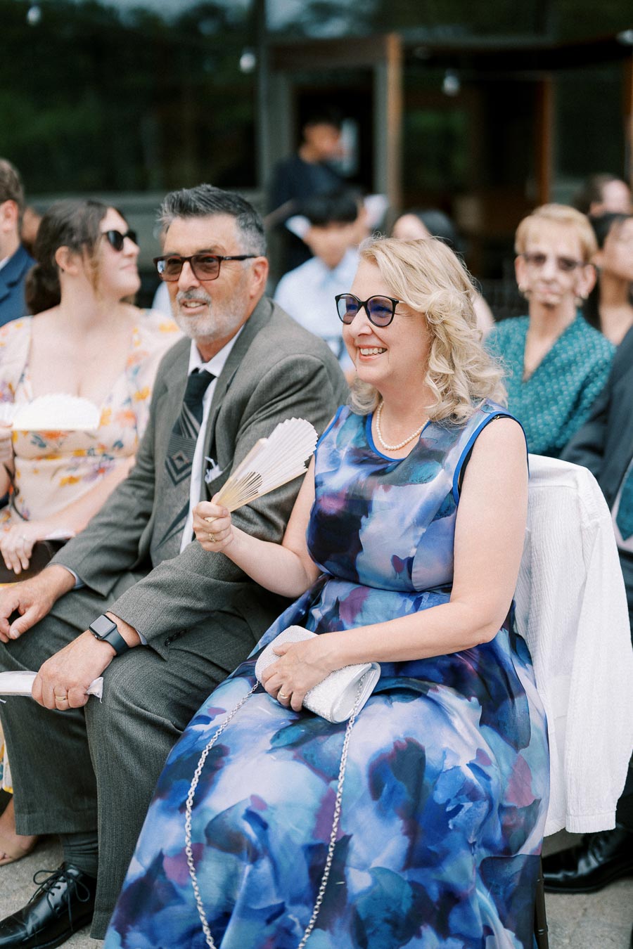 Guests at an outdoor wedding enjoying the ceremony, dressed in formal attire, with a woman in a floral dress holding a hand fan and smiling.