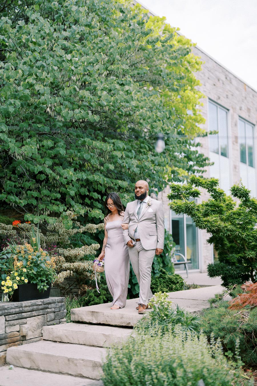 A couple in formal attire walking down stone steps in a lush garden setting, next to a modern building.