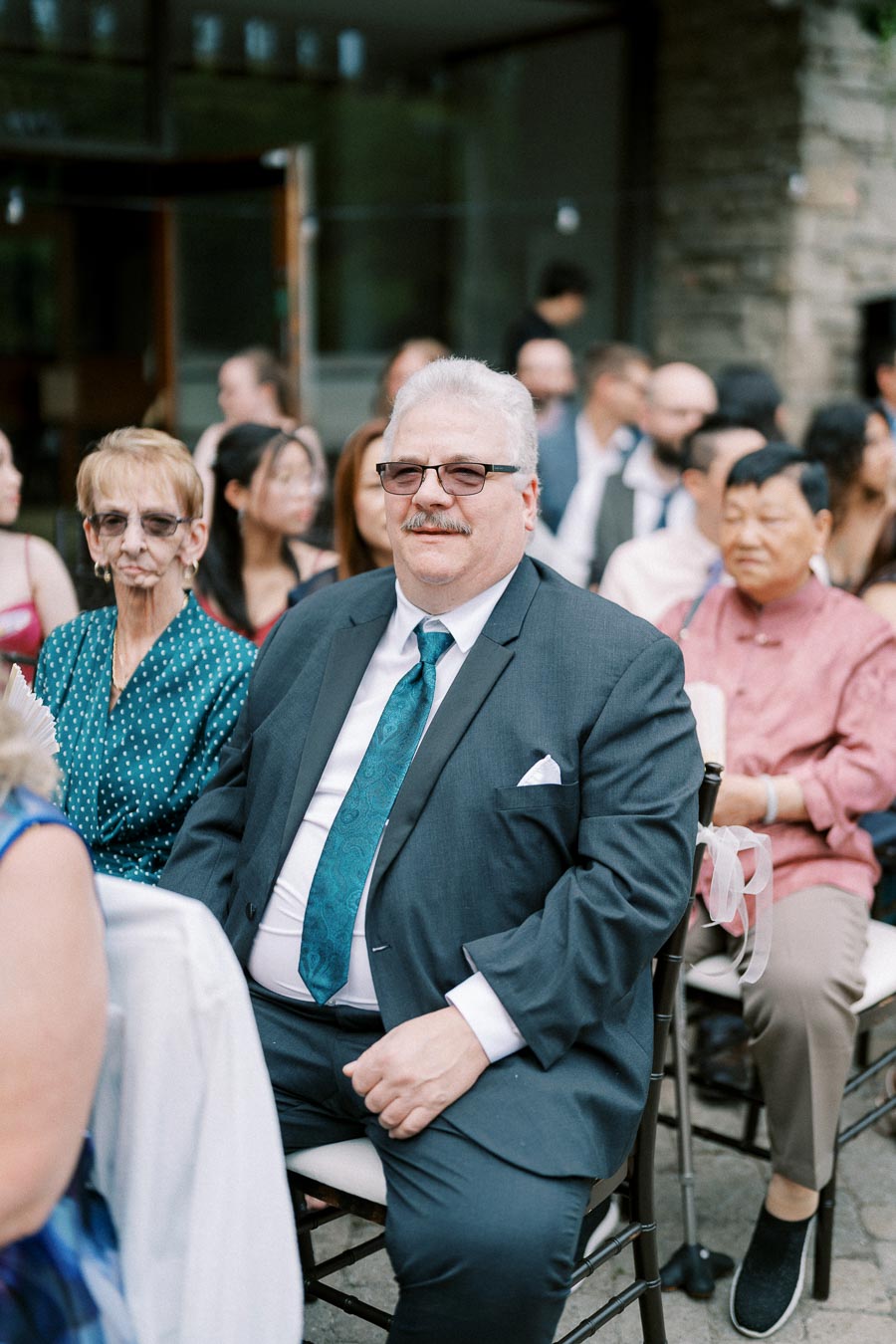 A group of elegantly dressed guests seated at an outdoor event, including a man in a suit with a teal tie and an older woman wearing glasses.