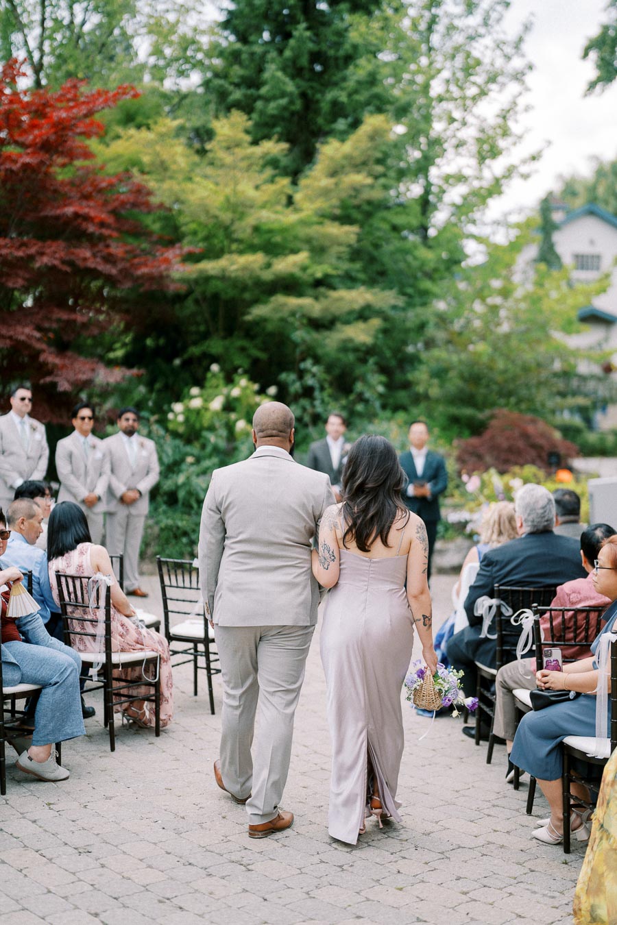 Outdoor wedding ceremony with a couple walking down the aisle surrounded by guests seated on both sides, lush greenery and trees in the background, with attendants in light suits standing at the front.