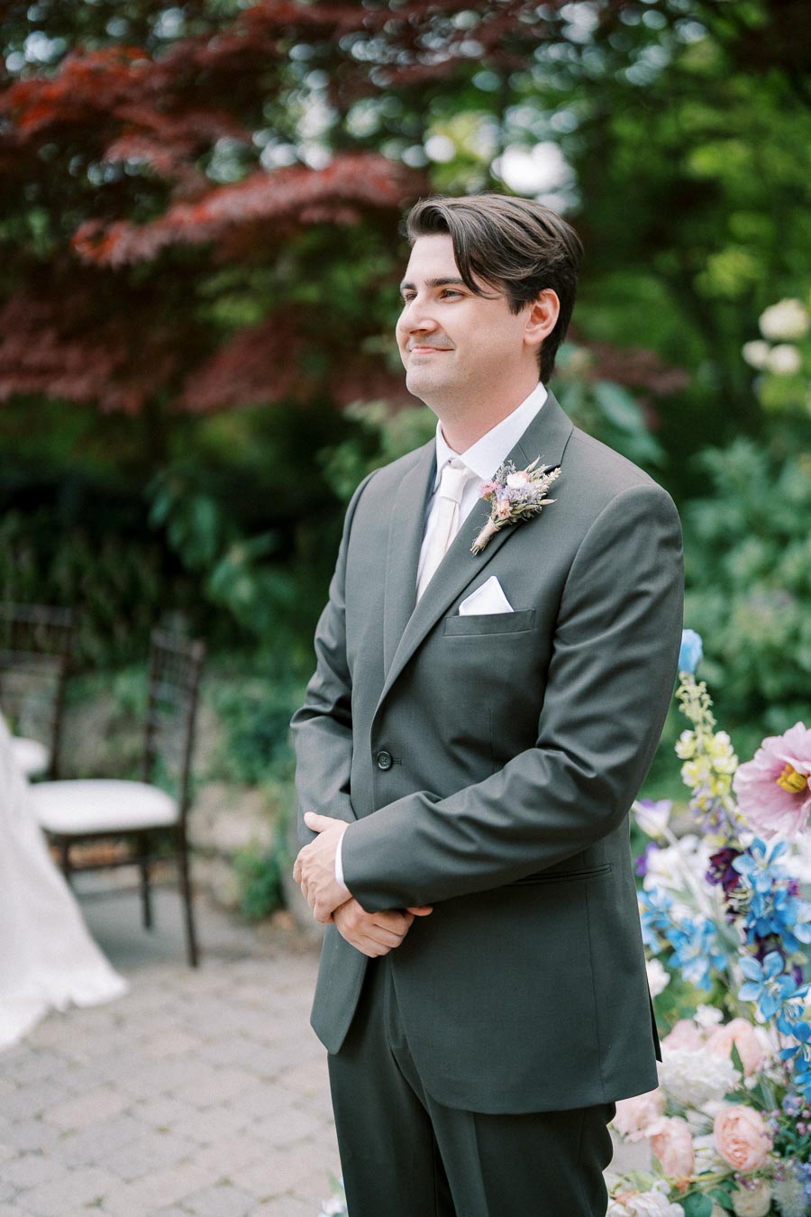 Groom in a dark suit smiling outdoors during a wedding ceremony, surrounded by vibrant floral arrangements and lush greenery.