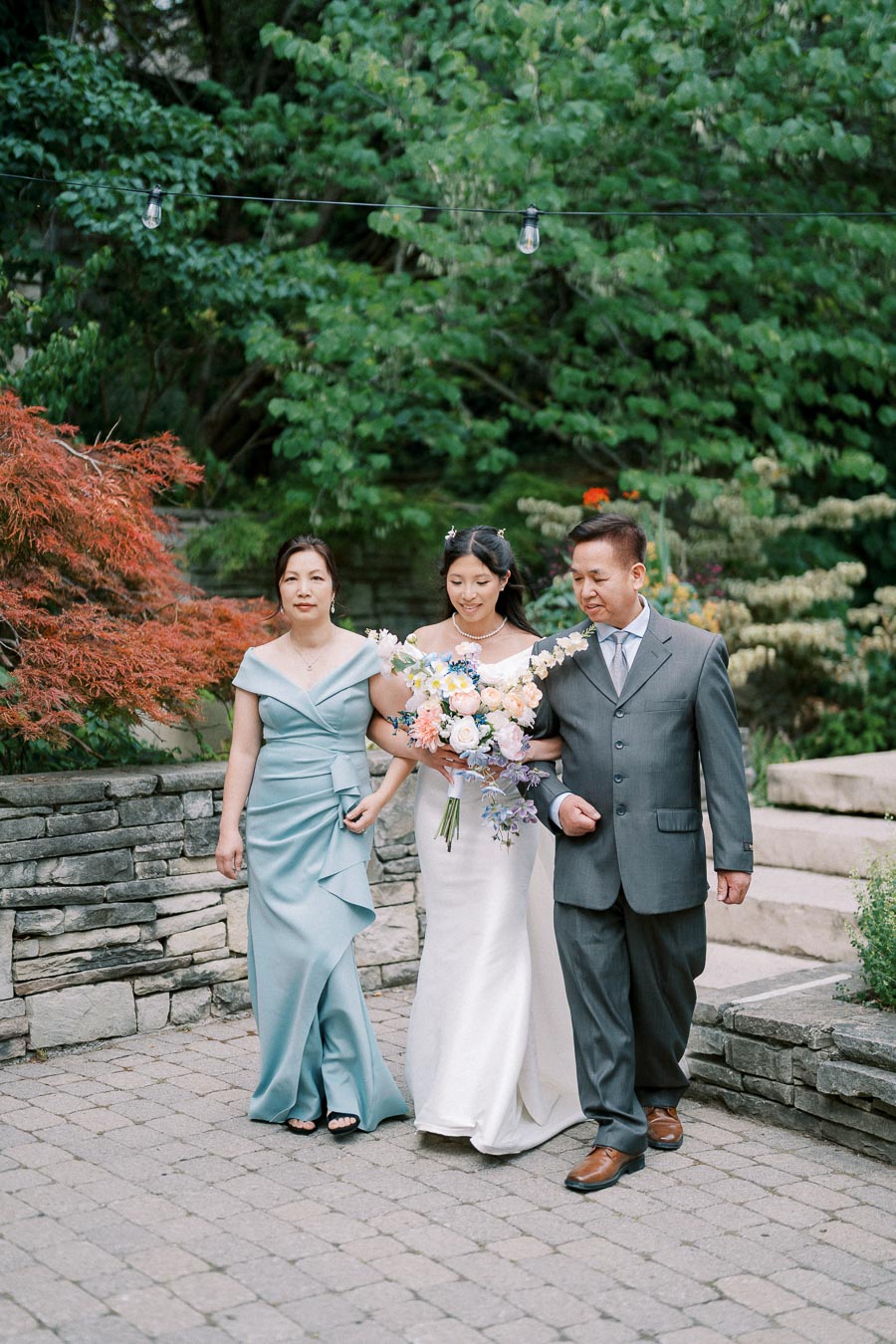 Bride walking with parents during outdoor wedding ceremony, holding a bouquet of pastel flowers, with lush greenery in the background.