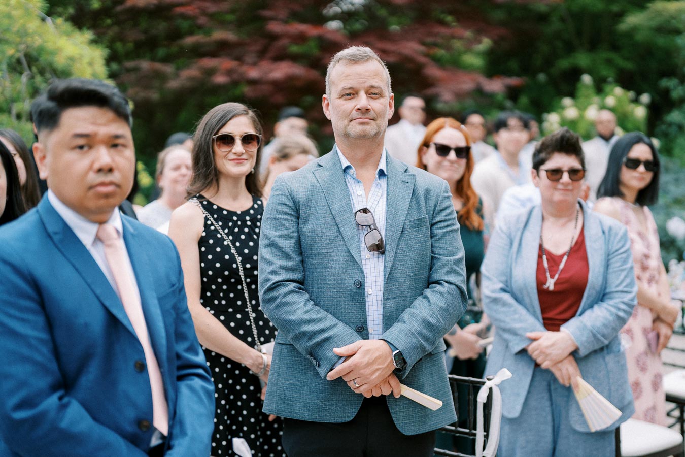 A group of well-dressed wedding guests standing outdoors, with greenery in the background, wearing sunglasses and holding paper fans on a sunny day.