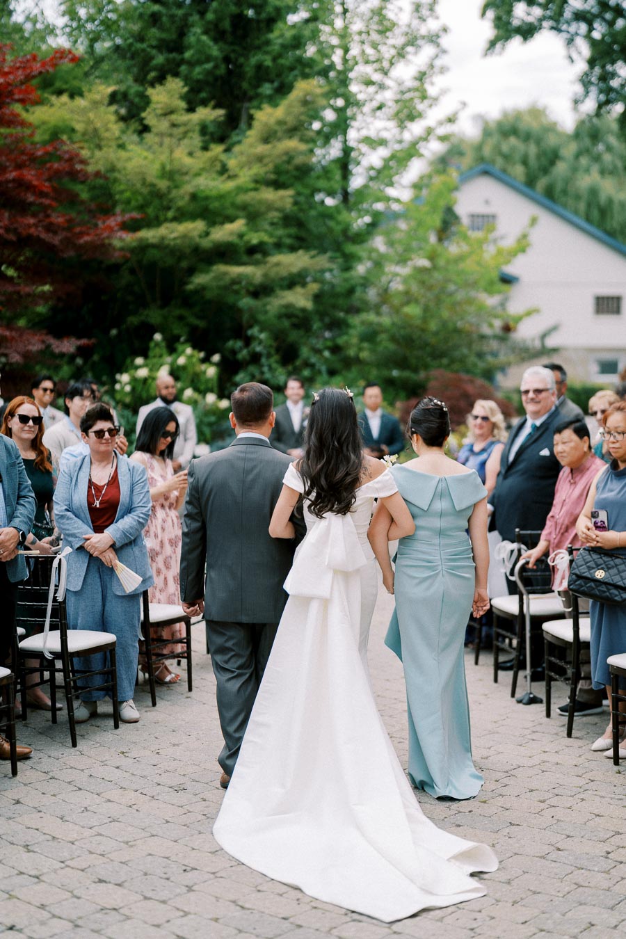 Wedding ceremony with bride in white gown walking down the aisle, accompanied by two family members, surrounded by guests at an outdoor venue with greenery and a rustic building backdrop.