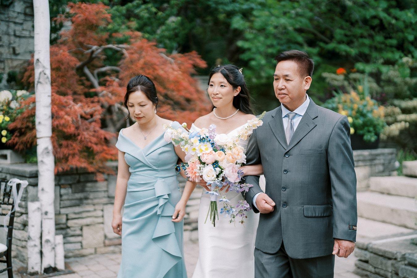 A bride in a white gown holding a colorful bouquet walks with her parents in elegant attire, set against a vibrant garden backdrop with red and green foliage.
