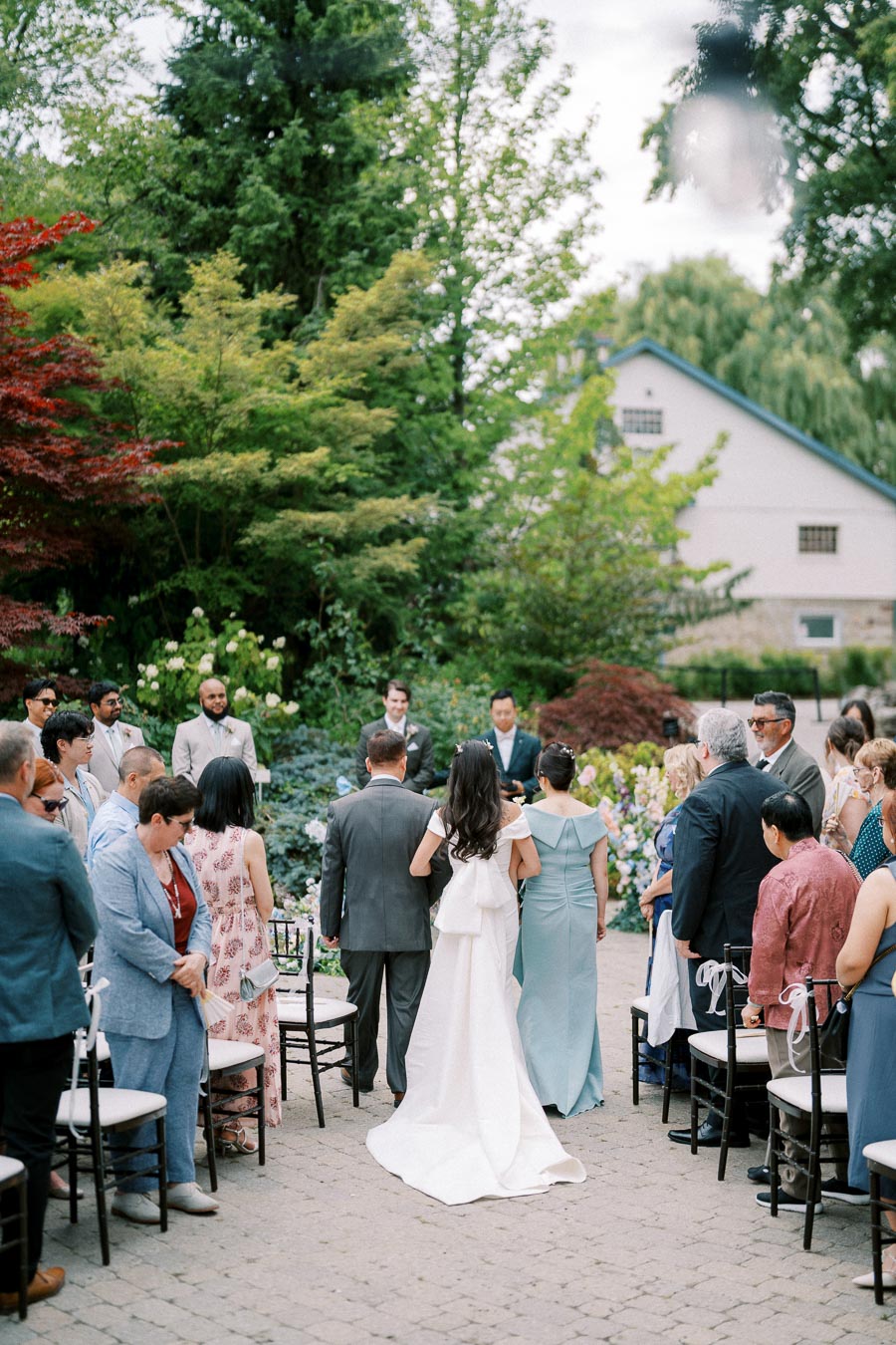 Outdoor wedding ceremony with a bride in a white dress walking with her father down the aisle, surrounded by seated guests in formal attire, set in a lush garden on a sunny day.