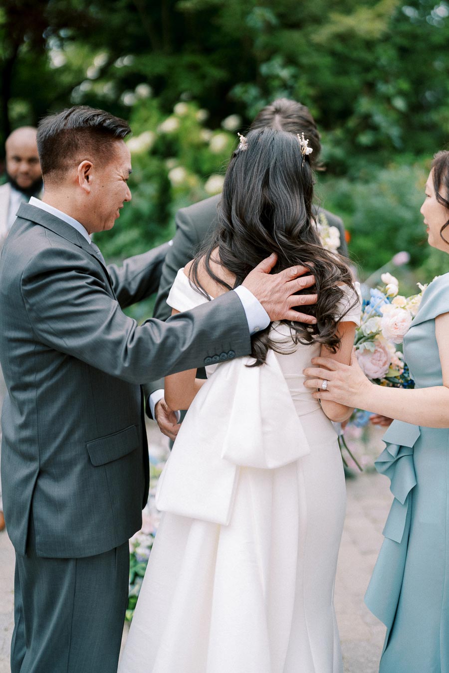 A bride in a white gown with a large bow is surrounded by family members in a lush garden setting, sharing a heartfelt moment before her wedding ceremony.