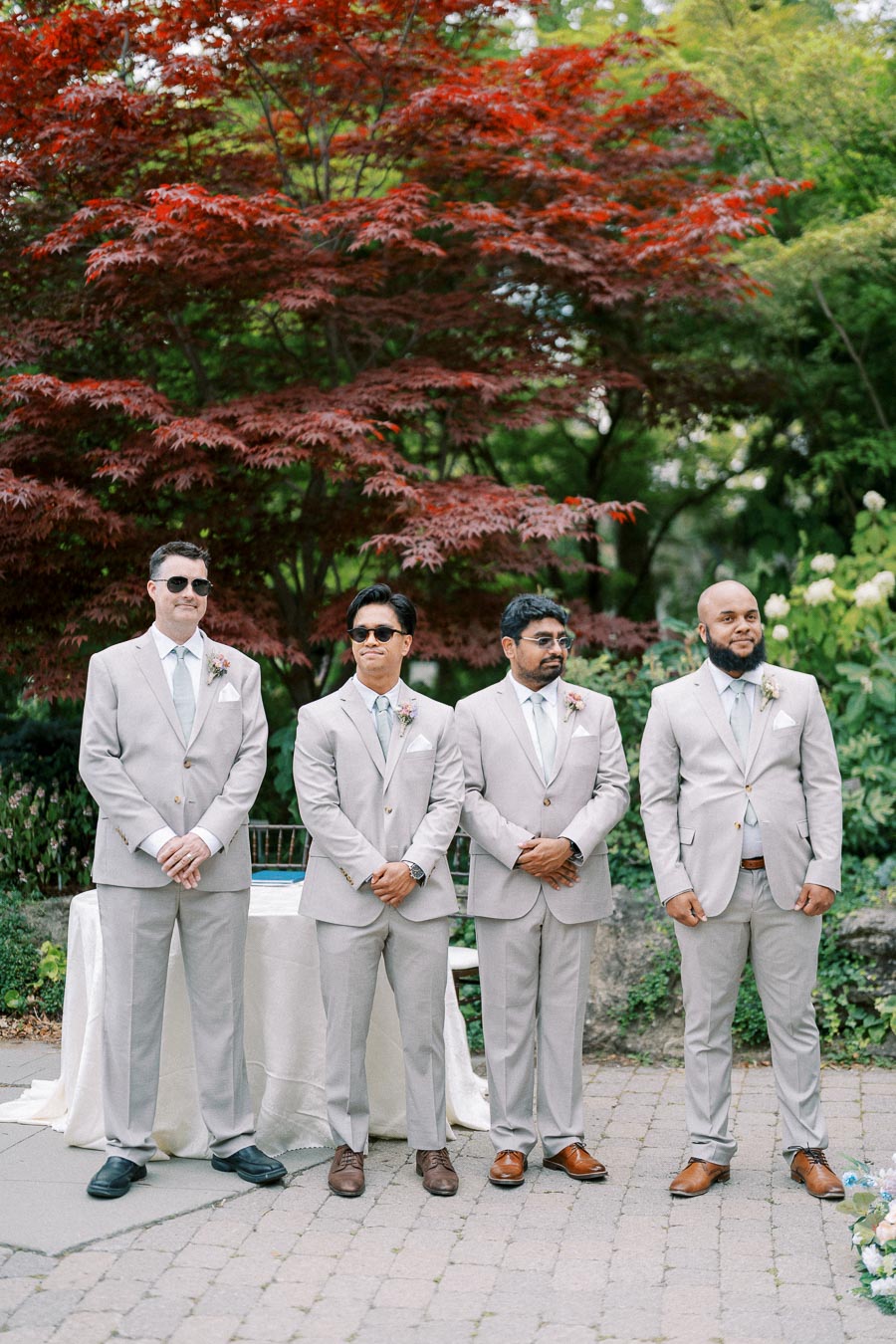 Four groomsmen in matching beige suits stand on a stone pathway, with vibrant greenery and red foliage in the background, highlighting a stylish outdoor wedding setting.