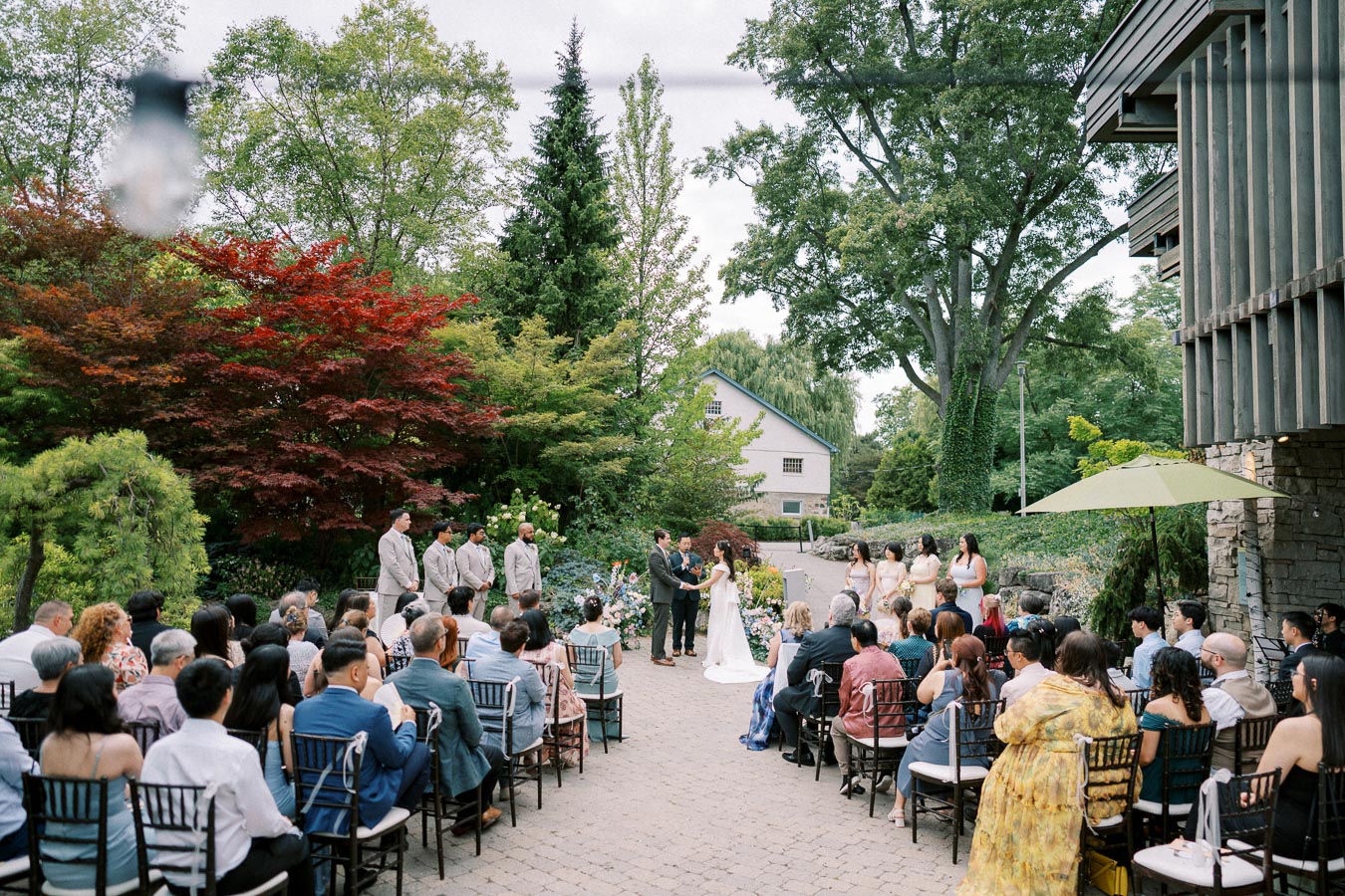 Outdoor wedding ceremony with guests seated on either side of the aisle. The couple stands at the altar surrounded by greenery and colorful flowers. Bridesmaids and groomsmen are positioned nearby, with lush trees and a rustic building in the background, creating a serene garden setting.