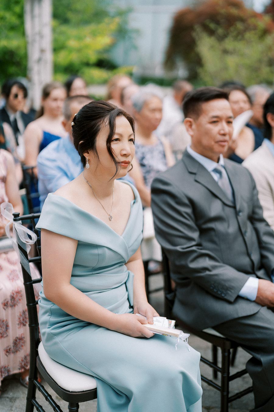 Guests seated at an outdoor wedding ceremony, woman in elegant light blue dress holding programs, surrounded by a crowd in formal attire, natural greenery in background.