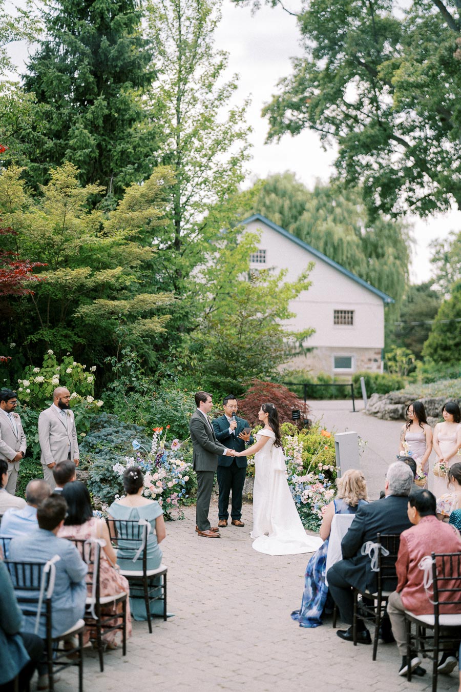 Outdoor garden wedding ceremony with a bride and groom exchanging vows, surrounded by floral arrangements and guests seated in rows, under a backdrop of lush green trees and a rustic building.