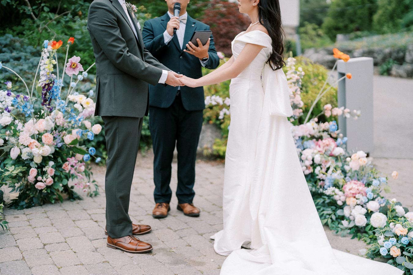 A bride and groom holding hands during an outdoor wedding ceremony, surrounded by colorful floral arrangements and an officiant speaking, creating a romantic and elegant atmosphere.