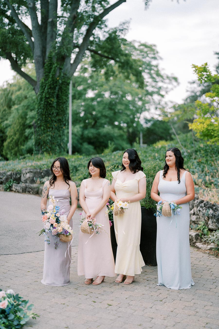 Four elegantly dressed bridesmaids in pastel gowns, standing outdoors with vibrant floral bouquets, surrounded by lush greenery and a stone pathway.