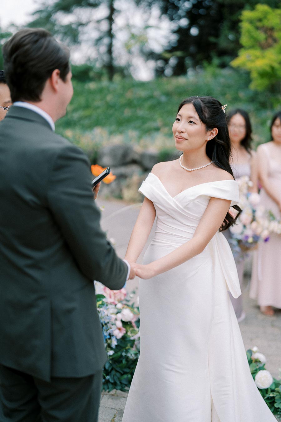 Bride and groom holding hands during outdoor wedding ceremony surrounded by greenery and bridesmaids in pastel dresses.