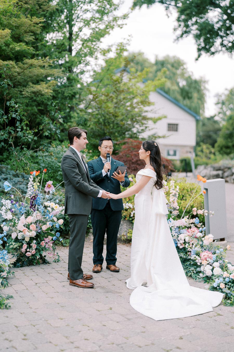 A bride and groom holding hands during their outdoor wedding ceremony, with an officiant standing between them, surrounded by colorful floral arrangements and lush green foliage.