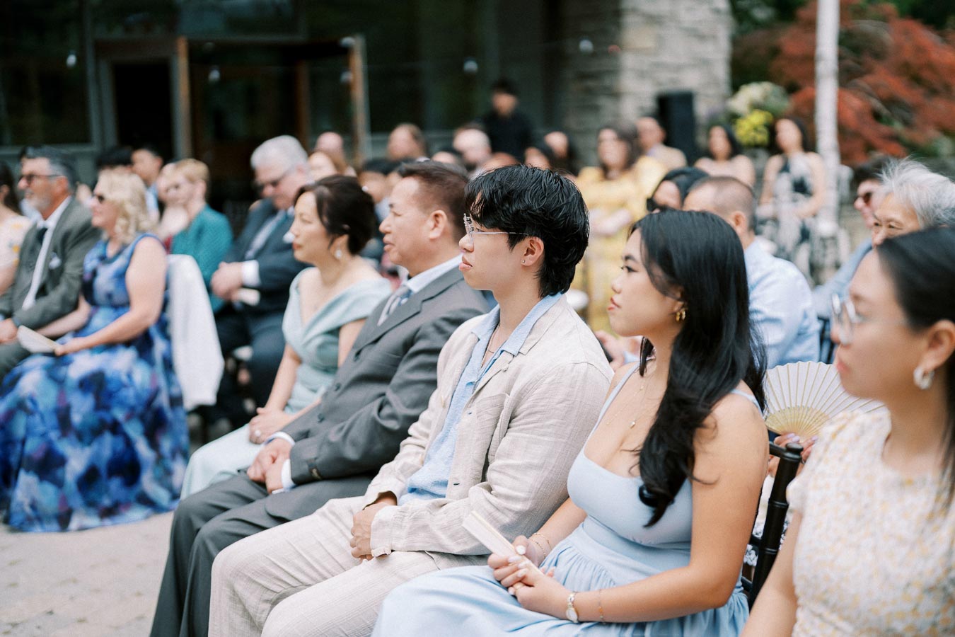 Guests attentively seated during an outdoor wedding ceremony, elegantly dressed in formal attire, with a focus on a couple wearing light blue outfits in the foreground.