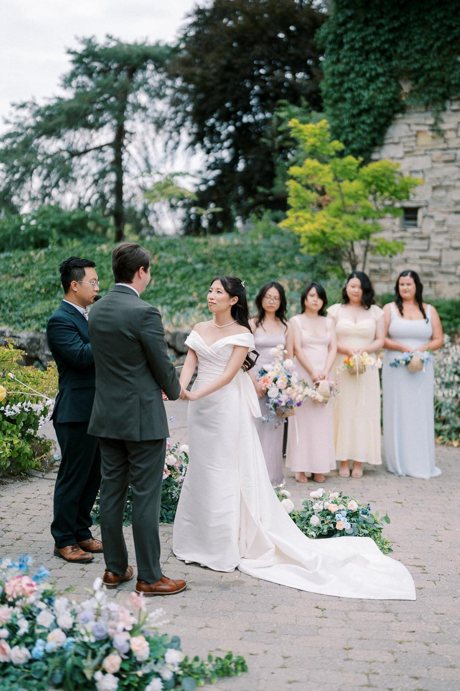 A bride and groom exchange vows outdoors during a wedding ceremony, surrounded by bridesmaids in pastel dresses and lush floral arrangements.