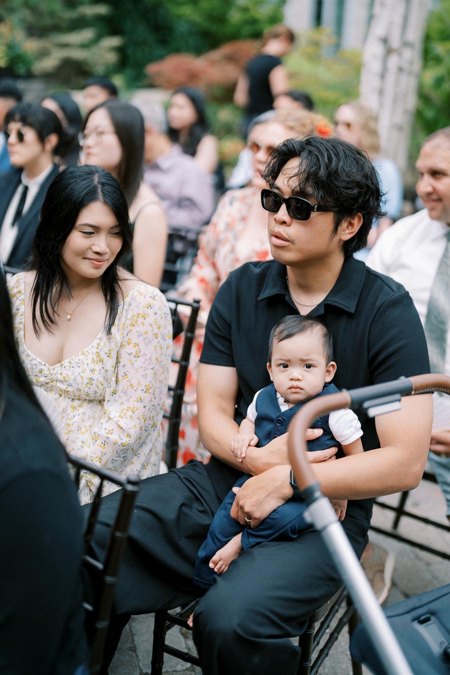 A man wearing sunglasses and holding a baby on his lap, seated among a diverse audience outdoors, with a woman beside him smiling, all appearing attentive and casually dressed.