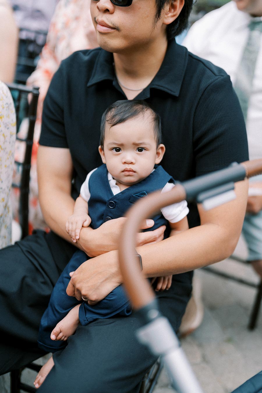 Father holding baby in formal attire at an outdoor event, both looking at the camera.