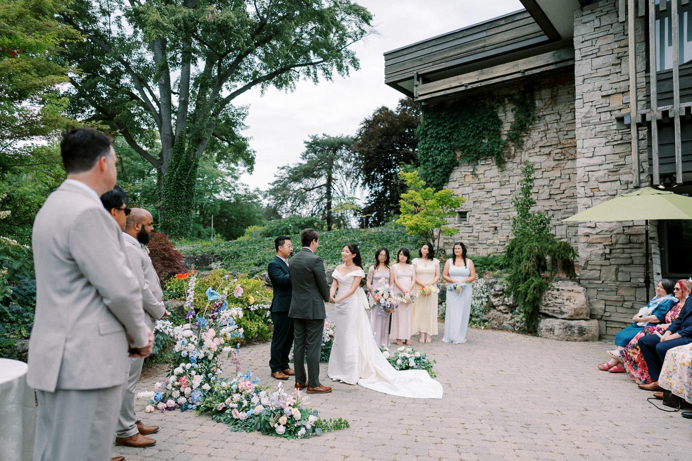 Outdoor wedding ceremony at a picturesque venue with lush greenery, a stone building backdrop, and floral arrangements. Bride and groom stand facing each other, surrounded by bridesmaids holding bouquets, while guests watch from the side.