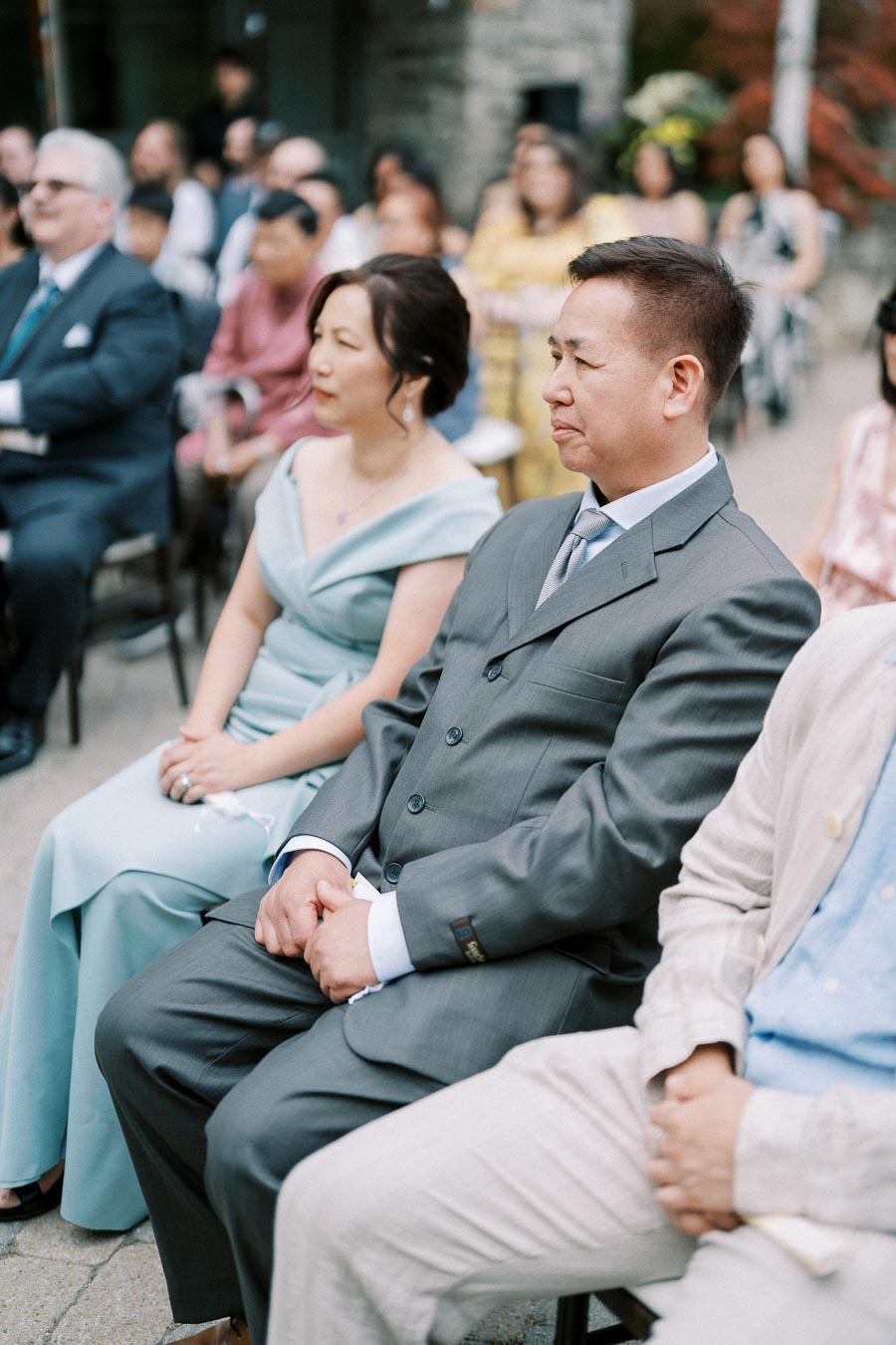 A close-up of a man in a gray suit and a woman in a light blue dress seated at an outdoor event, with other attendees and greenery in the background.