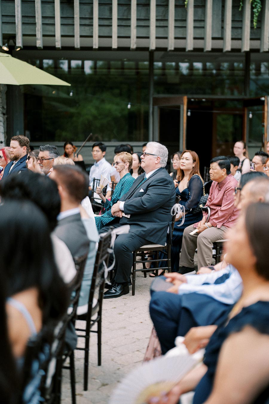 A group of people attentively seated during an outdoor event, with a mix of men and women in formal attire, under a partially covered structure with wooden beams.