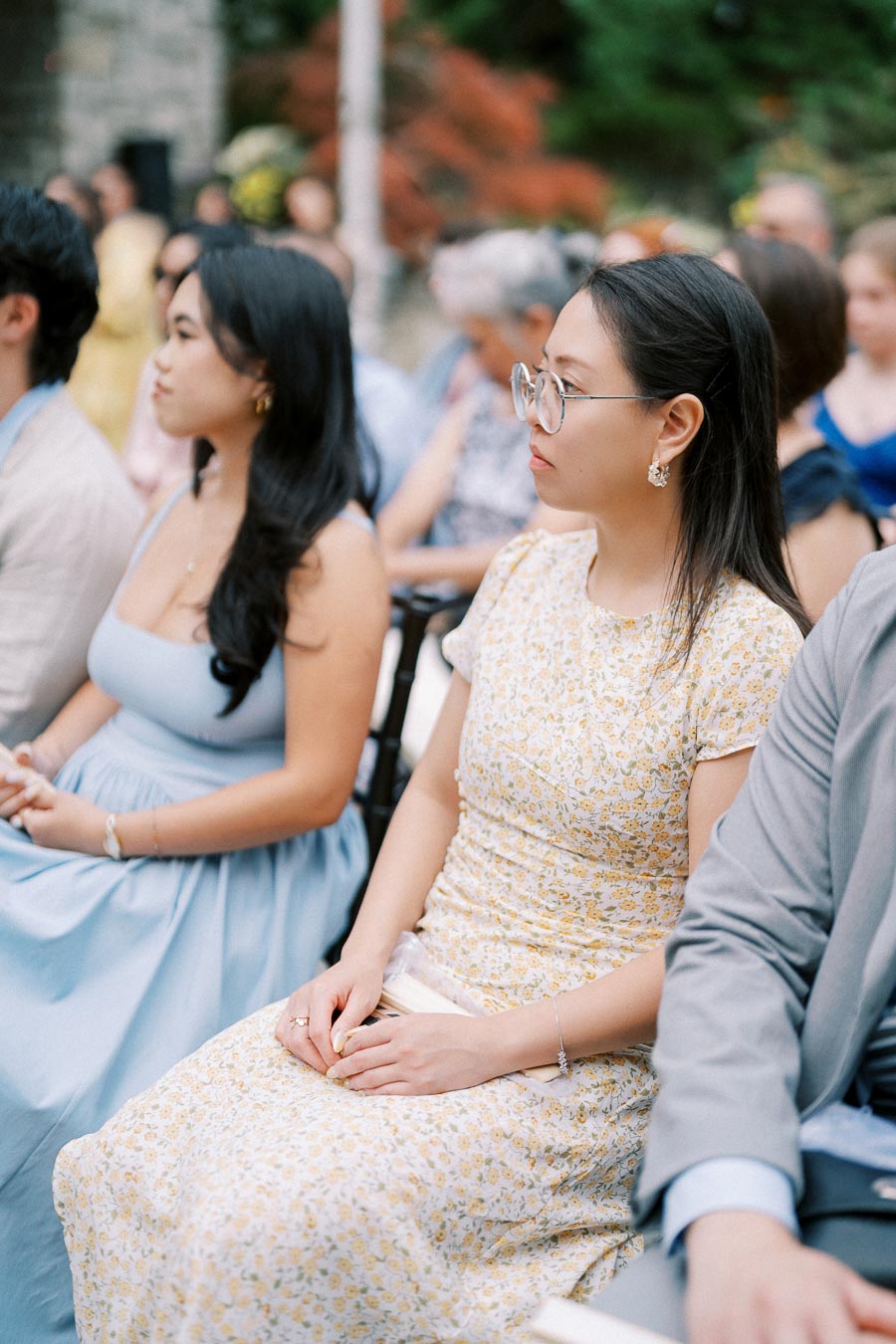 Outdoor event with attendees seated attentively, featuring a woman in a yellow floral dress and another in a light blue dress, surrounded by others in formal attire