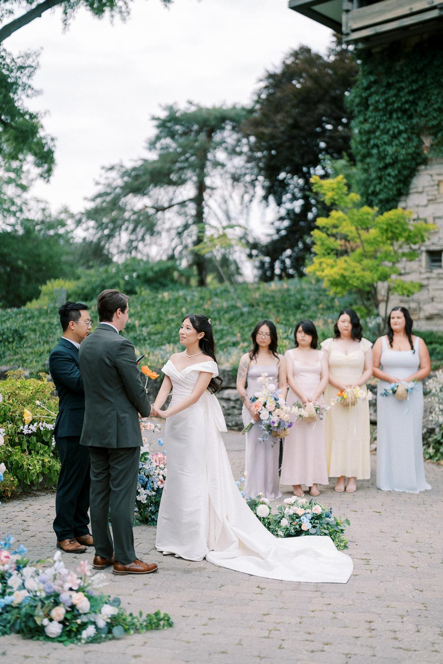 Outdoor wedding ceremony with bride in elegant white gown holding hands with groom, surrounded by bridesmaids in pastel dresses and floral arrangements, set in a lush garden backdrop.