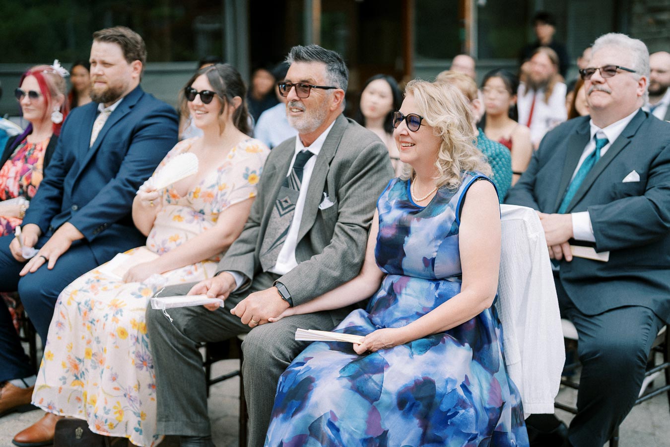A diverse group of well-dressed wedding guests sitting outdoors, smiling and wearing sunglasses, enjoying the ceremony on a sunny day.