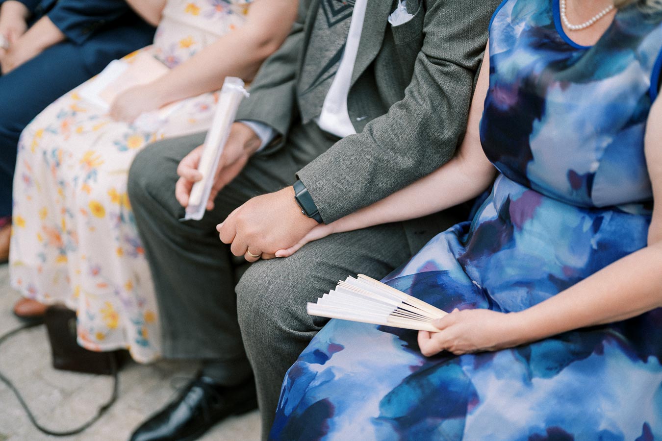 A group of formally dressed people seated, holding fans, with two individuals holding hands at an outdoor event.