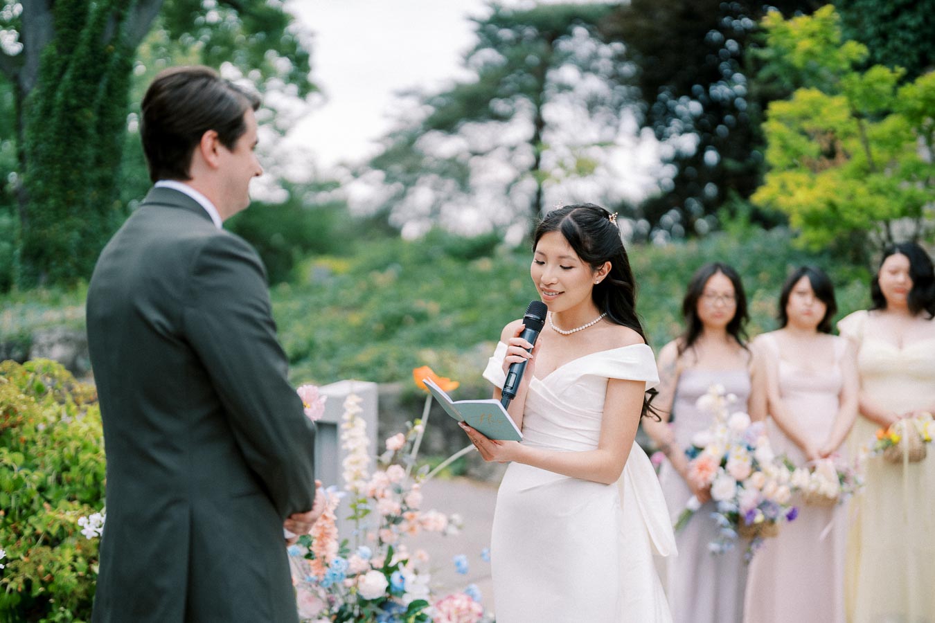 Bride reading vows to groom during outdoor wedding ceremony, surrounded by bridesmaids with colorful bouquets in a lush garden setting.