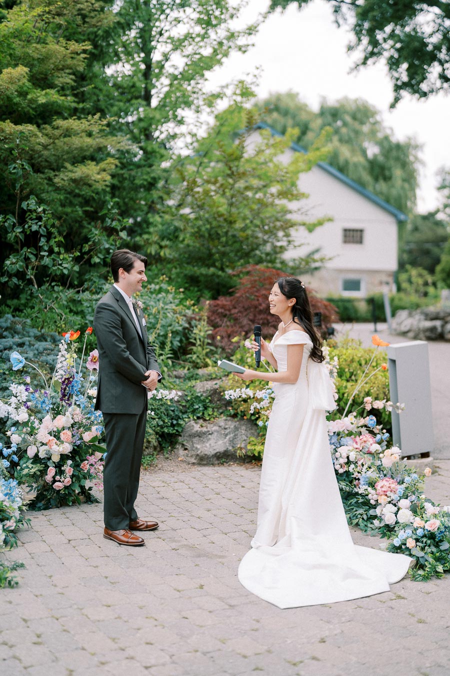 Bride and groom exchanging vows in a lush garden setting, surrounded by colorful floral arrangements, during an outdoor wedding ceremony.