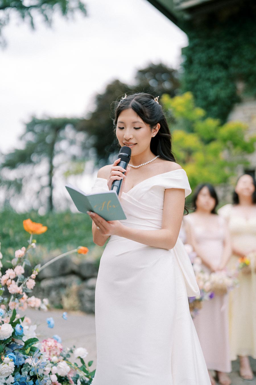 A woman in a white dress giving a speech with a microphone, holding a blue booklet, surrounded by colorful flowers and greenery outdoors.