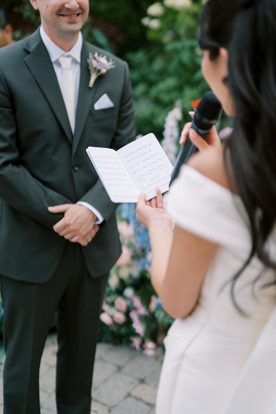 Bride reading vows to groom during outdoor wedding ceremony, groom smiling wearing dark suit with boutonnière, lush greenery and floral arrangements in background.