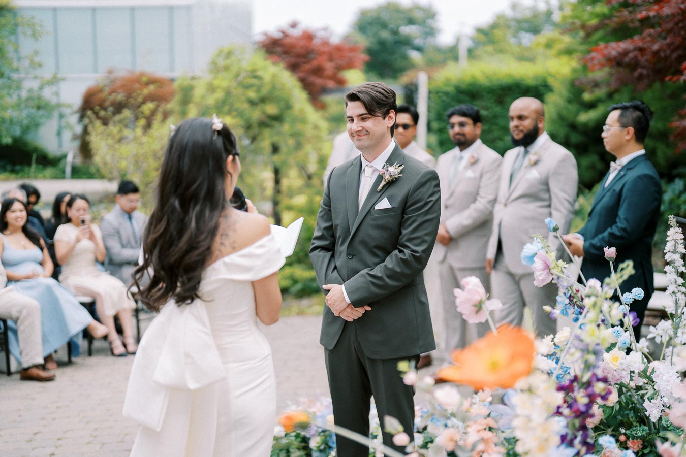 A bride and groom exchange vows during an outdoor wedding ceremony, surrounded by colorful flowers and guests. The groom is in a dark suit, while the bride wears an elegant off-the-shoulder white gown. Bridesmaids and groomsmen can be seen in the background in coordinated attire.
