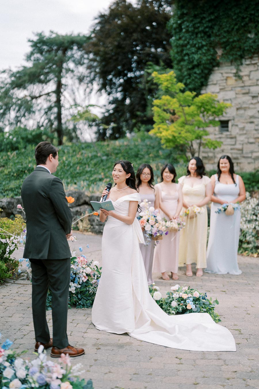Elegant outdoor wedding ceremony with a bride in a white gown reading vows to the groom, surrounded by bridesmaids in pastel dresses, with floral decorations and lush greenery in the background.
