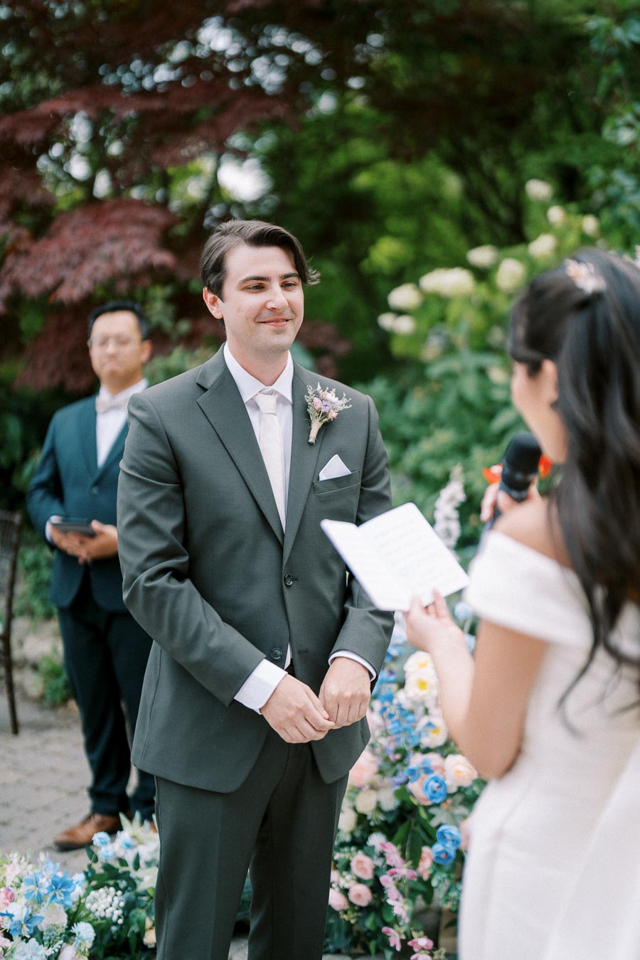 A groom in a dark suit stands facing the bride, who is holding a microphone and reading vows, during an outdoor wedding ceremony surrounded by lush greenery and colorful flowers.