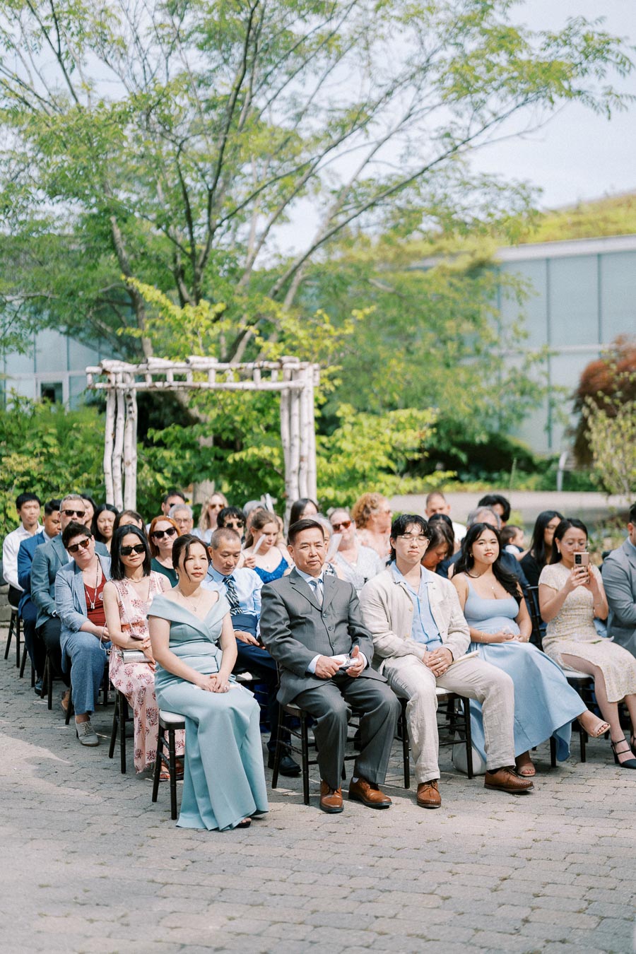 Outdoor wedding ceremony with guests seated under a lush green arbor, wearing formal attire on a sunny day.